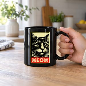 Person holding a glossy black ceramic mug with a bold cat portrait and red panel